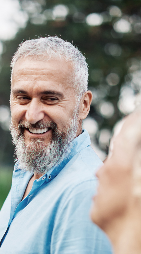 A bearded man smiling warmly while conversing outdoors, with greenery in the background. mobile