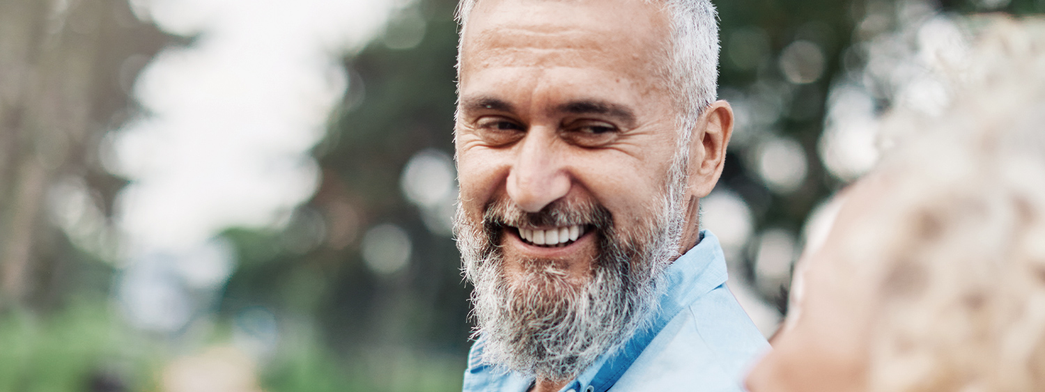 A bearded man smiling warmly while conversing outdoors, with greenery in the background. mobile