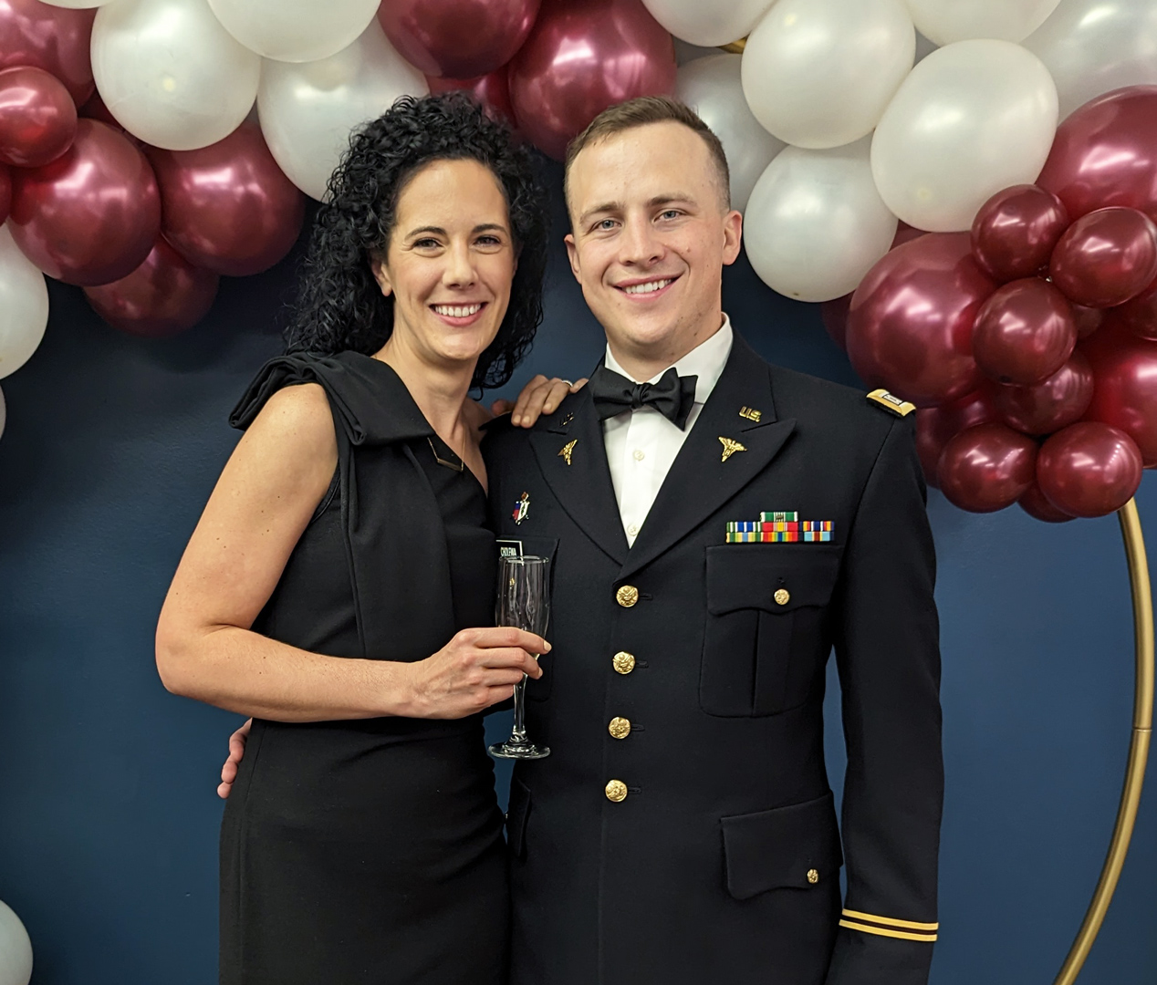 Dr. Cholewa, dressed in a formal military uniform, and a woman in a black dress smiling together in front of a balloon arch with maroon and white balloons.