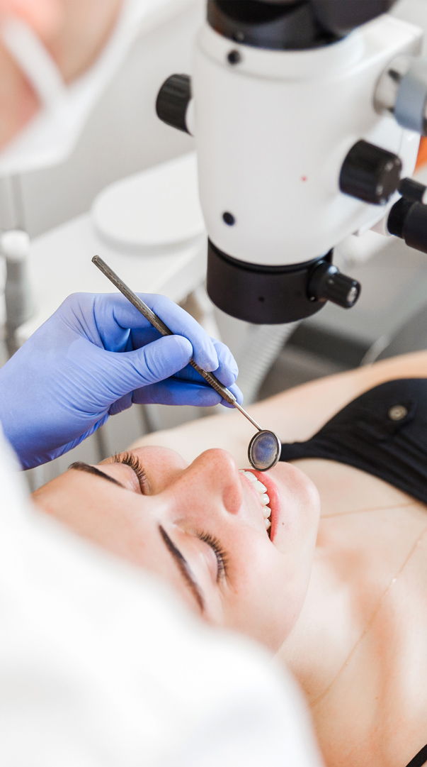 A patient smiling while undergoing a dental examination, with a dentist's gloved hand holding a dental mirror near the patient's mouth under a magnification device.